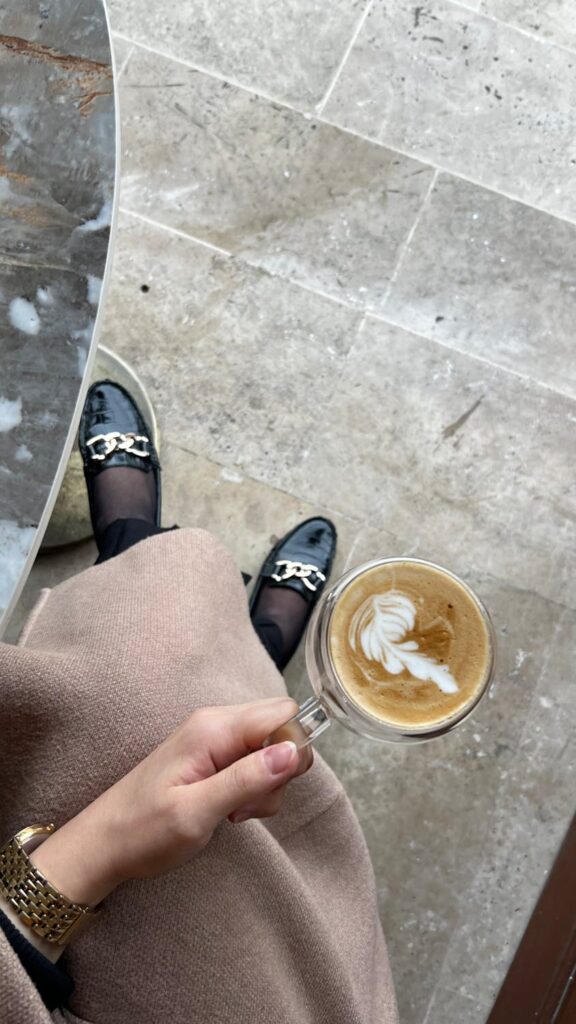 Person sitting at table with coffee in hand looking down at shoes. 